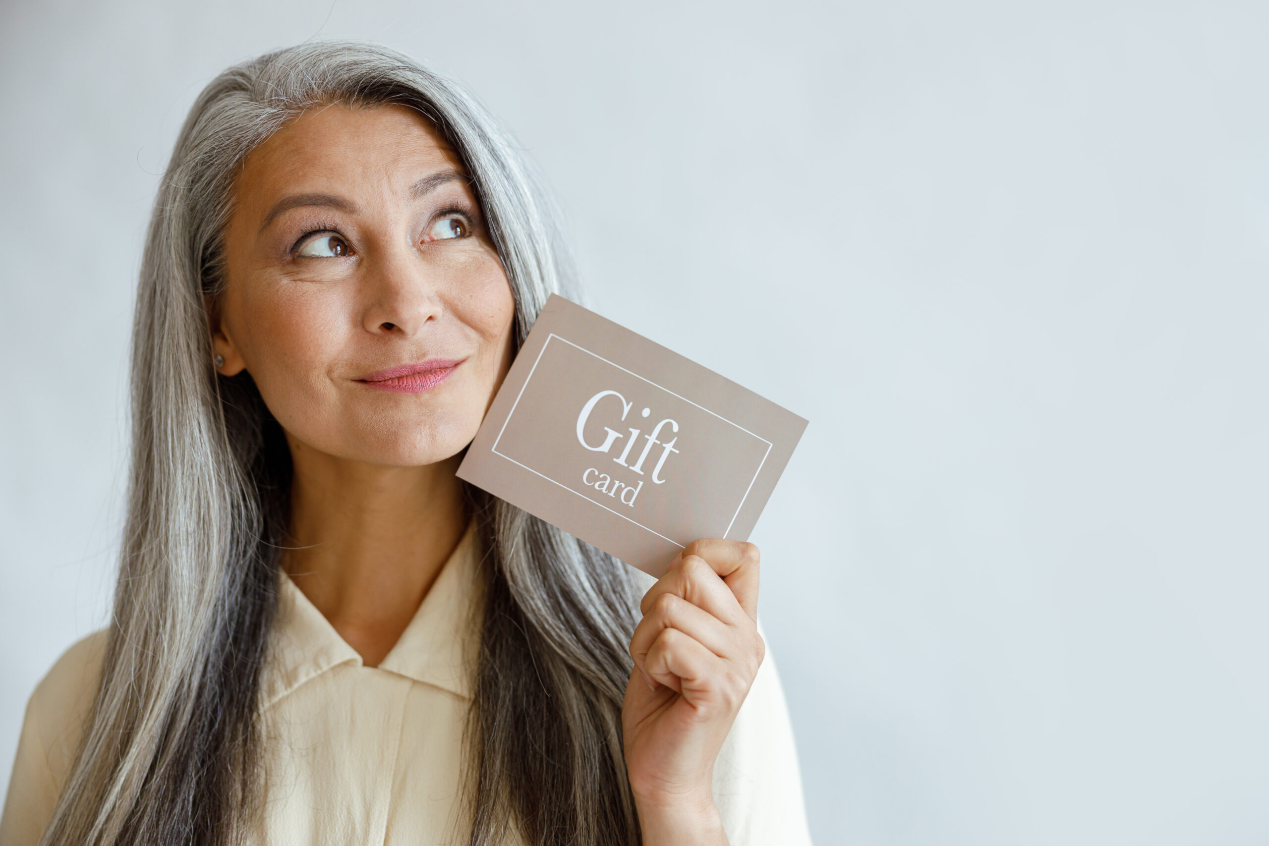 Dreamful grey haired Asian woman holds gift card near face in studio